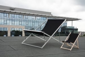 A lounge chair alongside a giant lounge chair among a backdrop of office buildings.