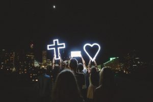 A group of people gathered in the city with neon signs of a cross and heart lit and held up above the crowd.