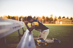 An American football player kneeling in prayer on a field bench.