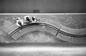 A black and white arial photo of people sitting on a bench that resembles music staves in contrast to the straight sidewalk.