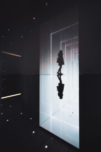 A person walking from a dark room into light, with their reflection following them on the floor.