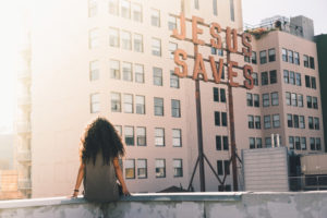 A person sitting on the edge of a roof facing a sign that reads 'Jesus Saves'.