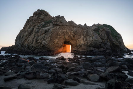 The sunset breaking through a hole in a rock formation along the coastline.