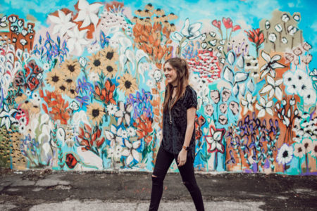 A woman smiling in front of a wall of graffiti flowers.